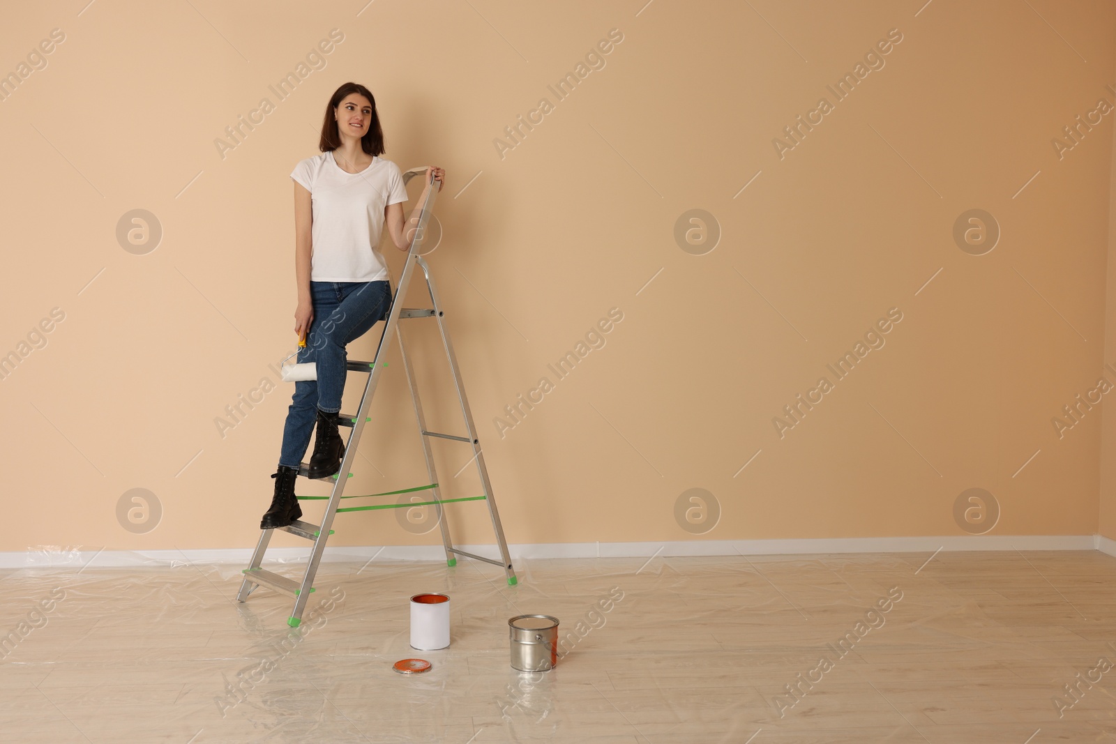 Photo of Young woman with roller on metal stepladder indoors. Space for text