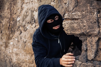 Photo of Man in mask behind stone wall watching someone outdoors. Criminal activity
