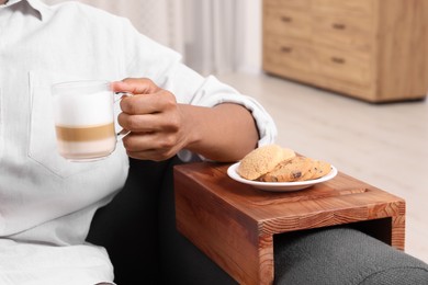 Cookies on sofa armrest wooden table. Man holding cup of coffee at home, closeup