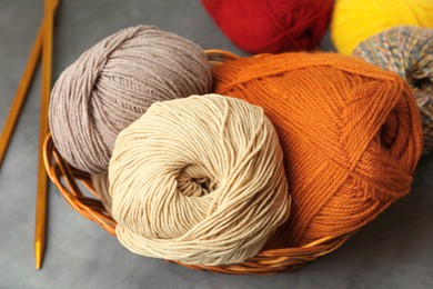 Photo of Different balls of woolen knitting yarns in wicker basket and needles on grey background, closeup