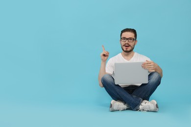 Emotional man sitting with laptop and pointing at something on light blue background. Space for text