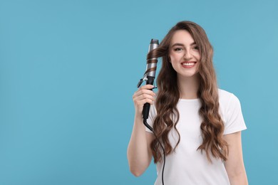 Beautiful young woman using curling hair iron on light blue background, space for text