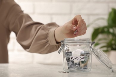 Photo of Pension savings. Woman putting coin into glass jar with money at light table, closeup