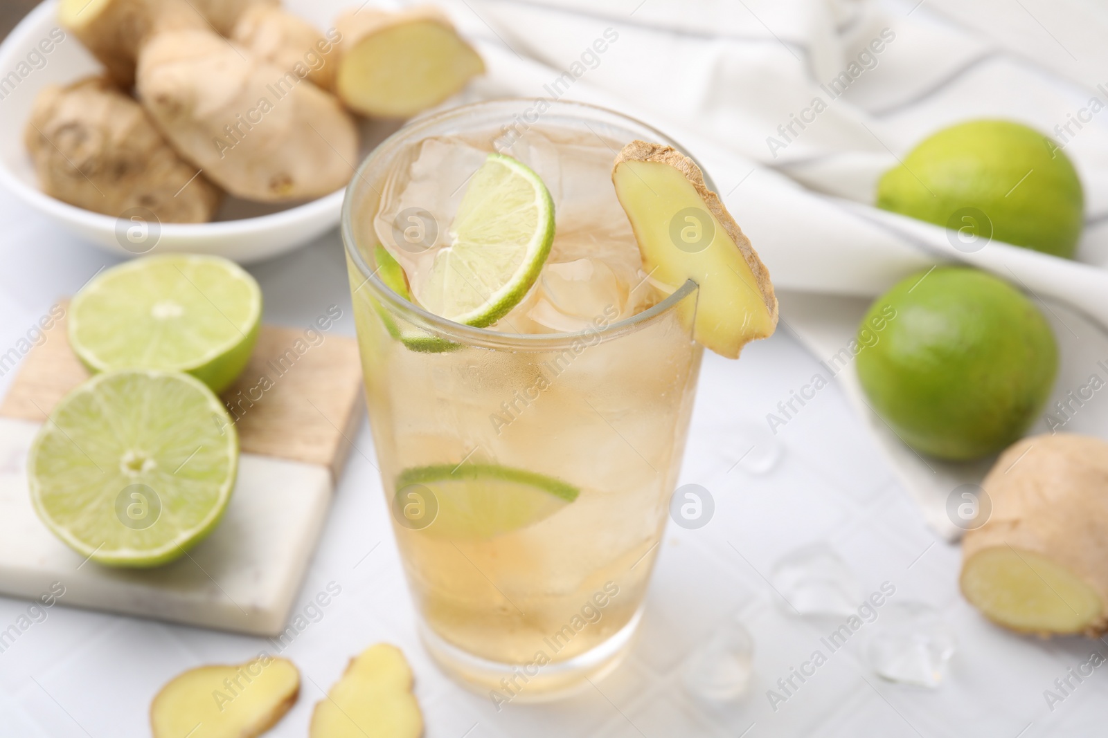 Photo of Glass of tasty ginger ale with ice cubes and ingredients on white tiled table, closeup