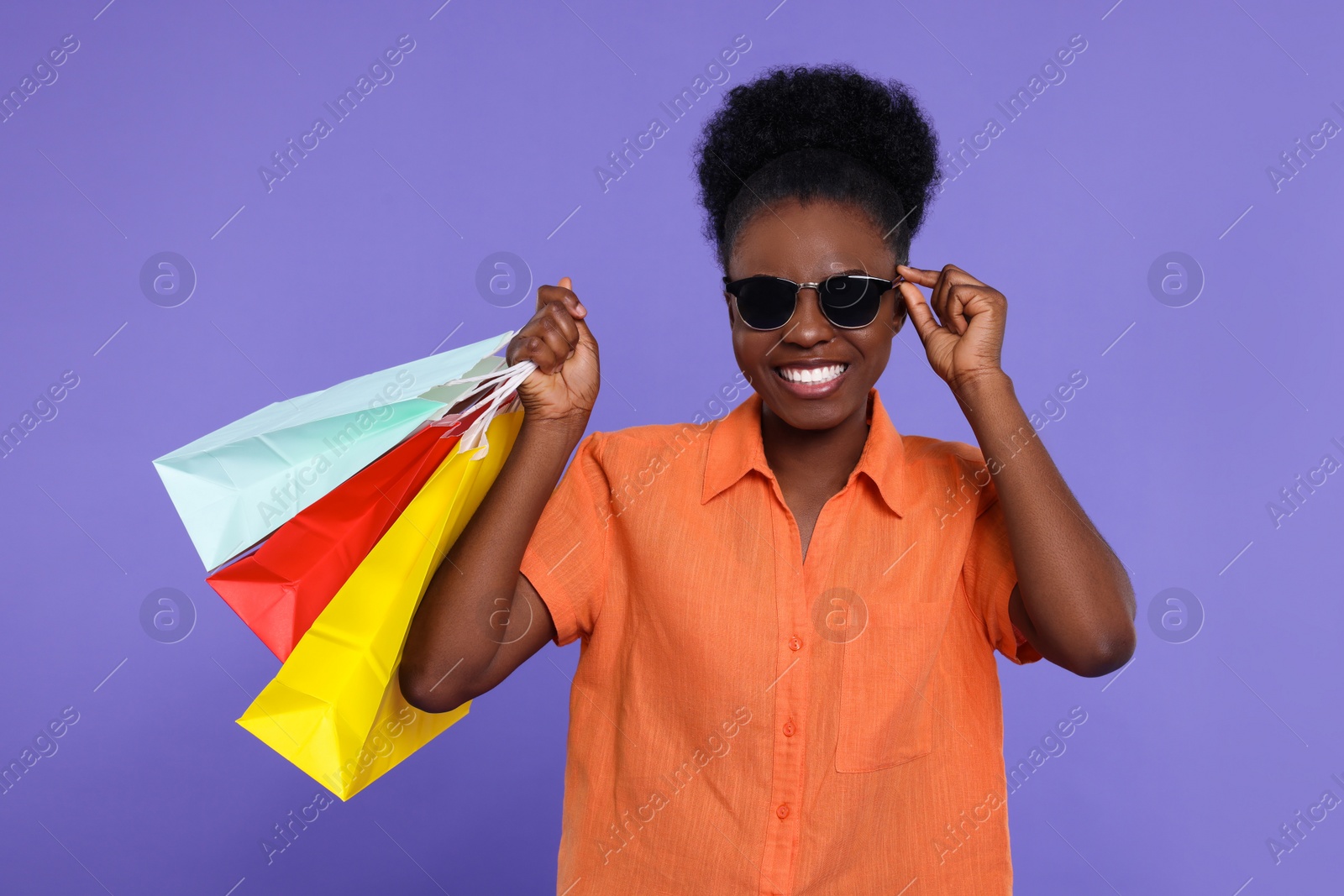 Photo of Happy young woman in stylish sunglasses with shopping bags on purple background