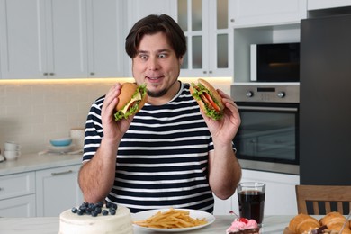 Hungry overweight man with tasty burgers at table in kitchen