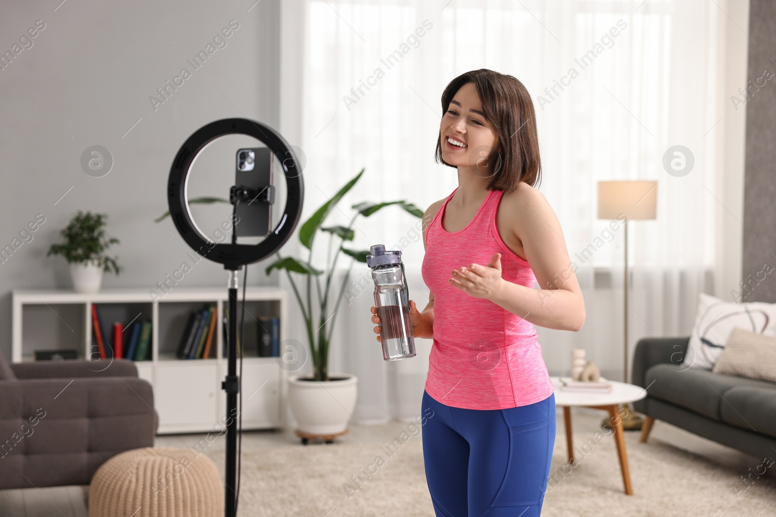 Photo of Happy sports blogger with bottle streaming online fitness lesson with smartphone at home