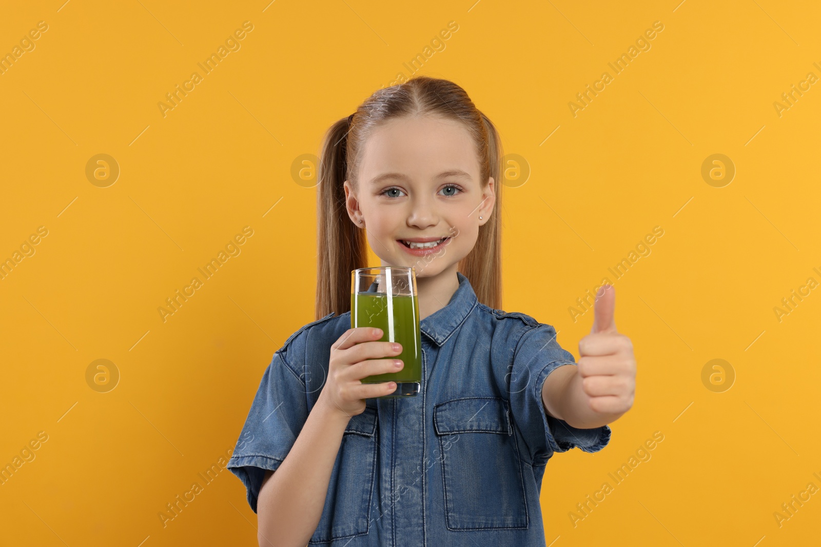 Photo of Cute little girl with glass of fresh juice showing thumbs up on orange background