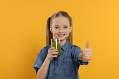 Photo of Cute little girl with glass of fresh juice showing thumbs up on orange background