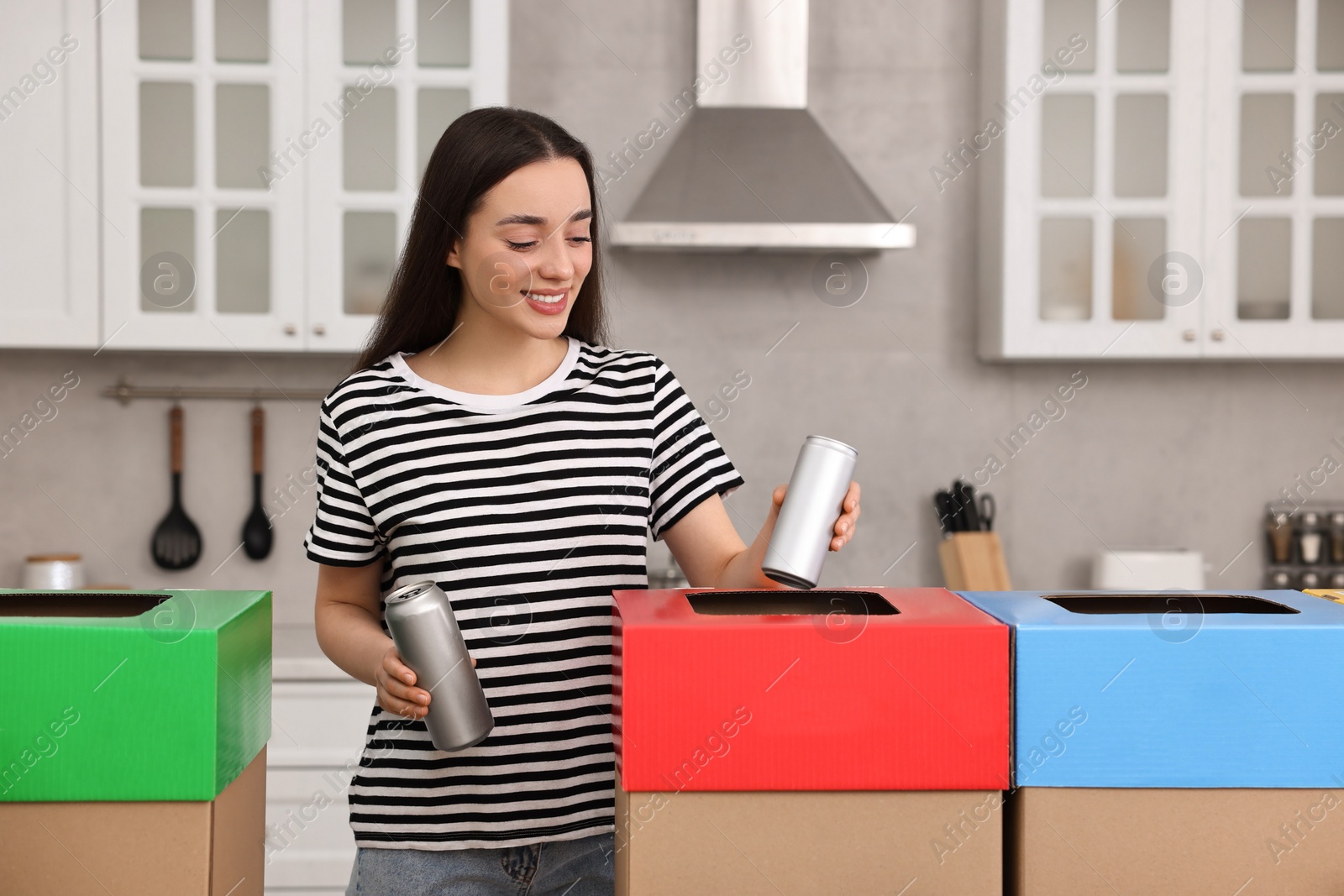 Photo of Garbage sorting. Smiling woman throwing metal can into cardboard box in kitchen