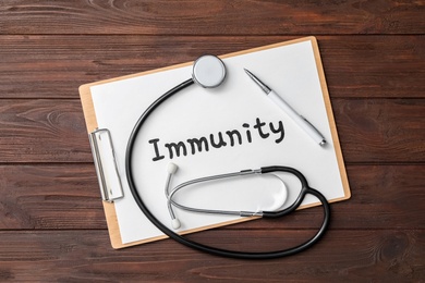 Clipboard with word Immunity and stethoscope on wooden table, top view