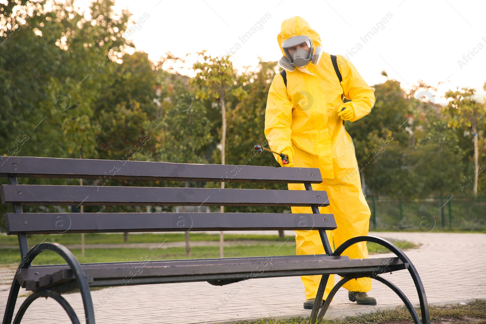 Photo of Person in hazmat suit disinfecting bench in park with sprayer. Surface treatment during coronavirus pandemic