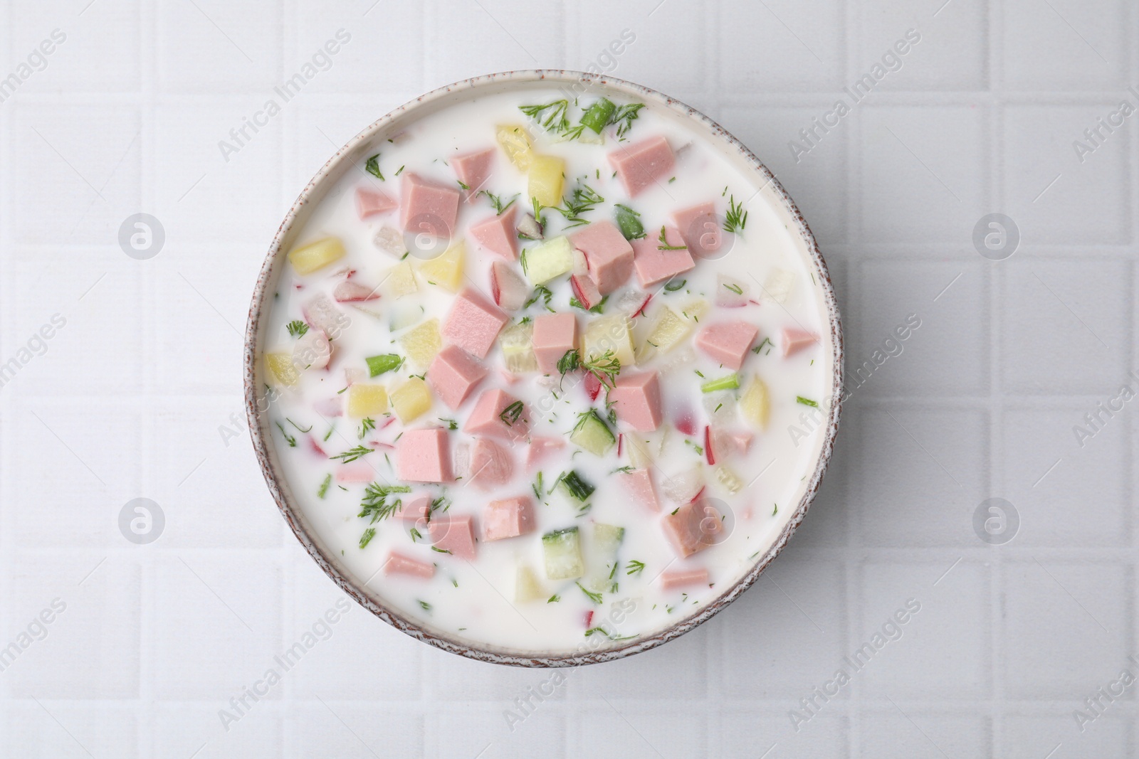 Photo of Delicious cold summer soup (okroshka) with boiled sausage in bowl on white tiled table, top view
