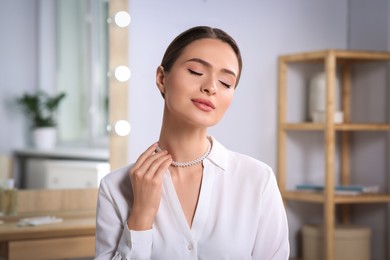 Photo of Young woman trying on elegant pearl necklace indoors