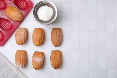 Delicious madeleine cookies, sieve with powdered sugar and baking mold on white tiled table, flat lay. Space for text