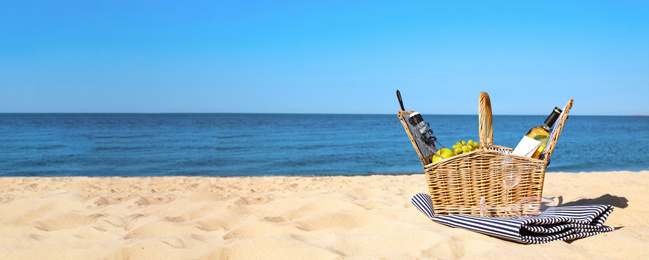 Wicker picnic basket with products and blanket on sunny beach, space for text. Banner design