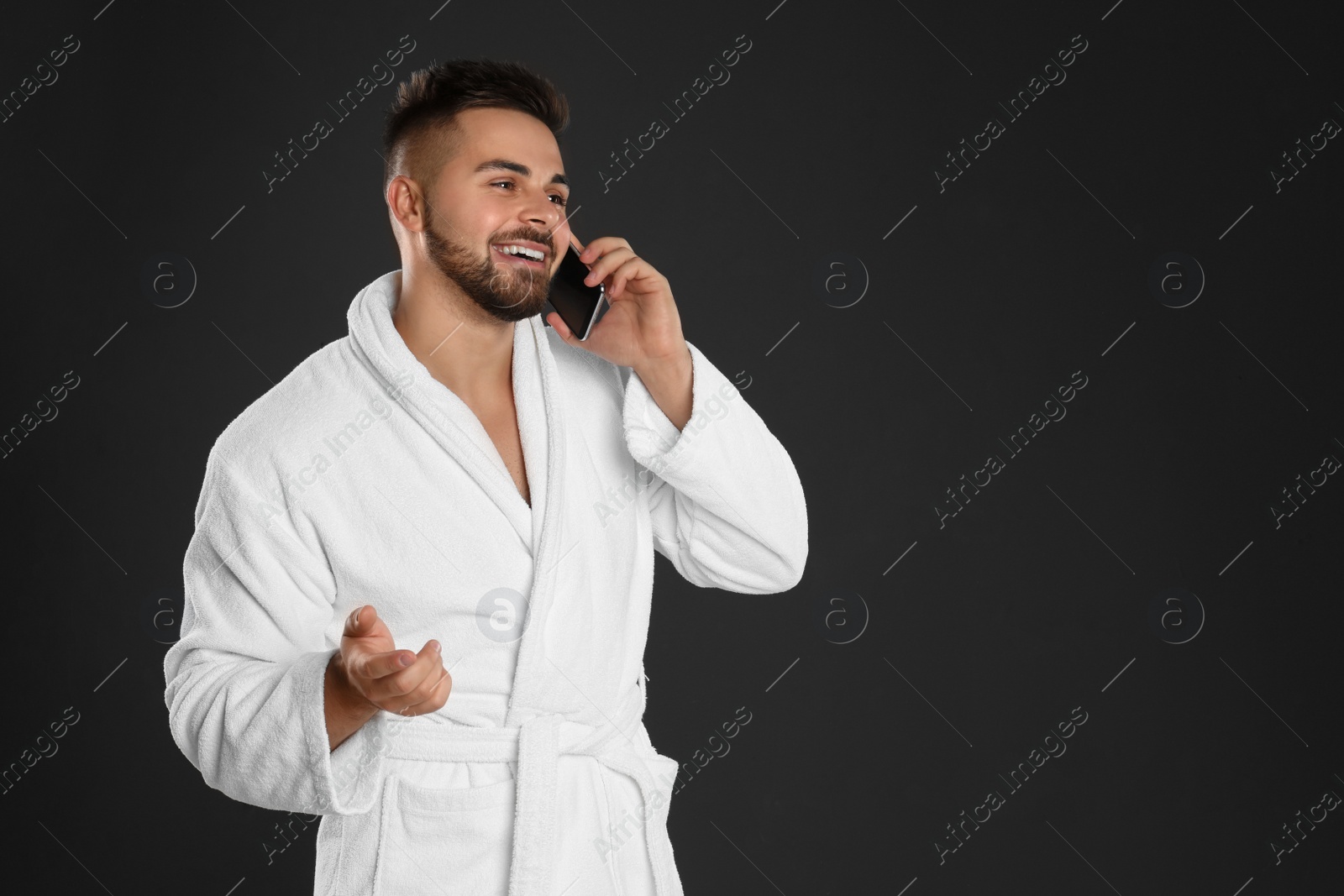 Photo of Young man in bathrobe talking on mobile phone against black background. Space for text