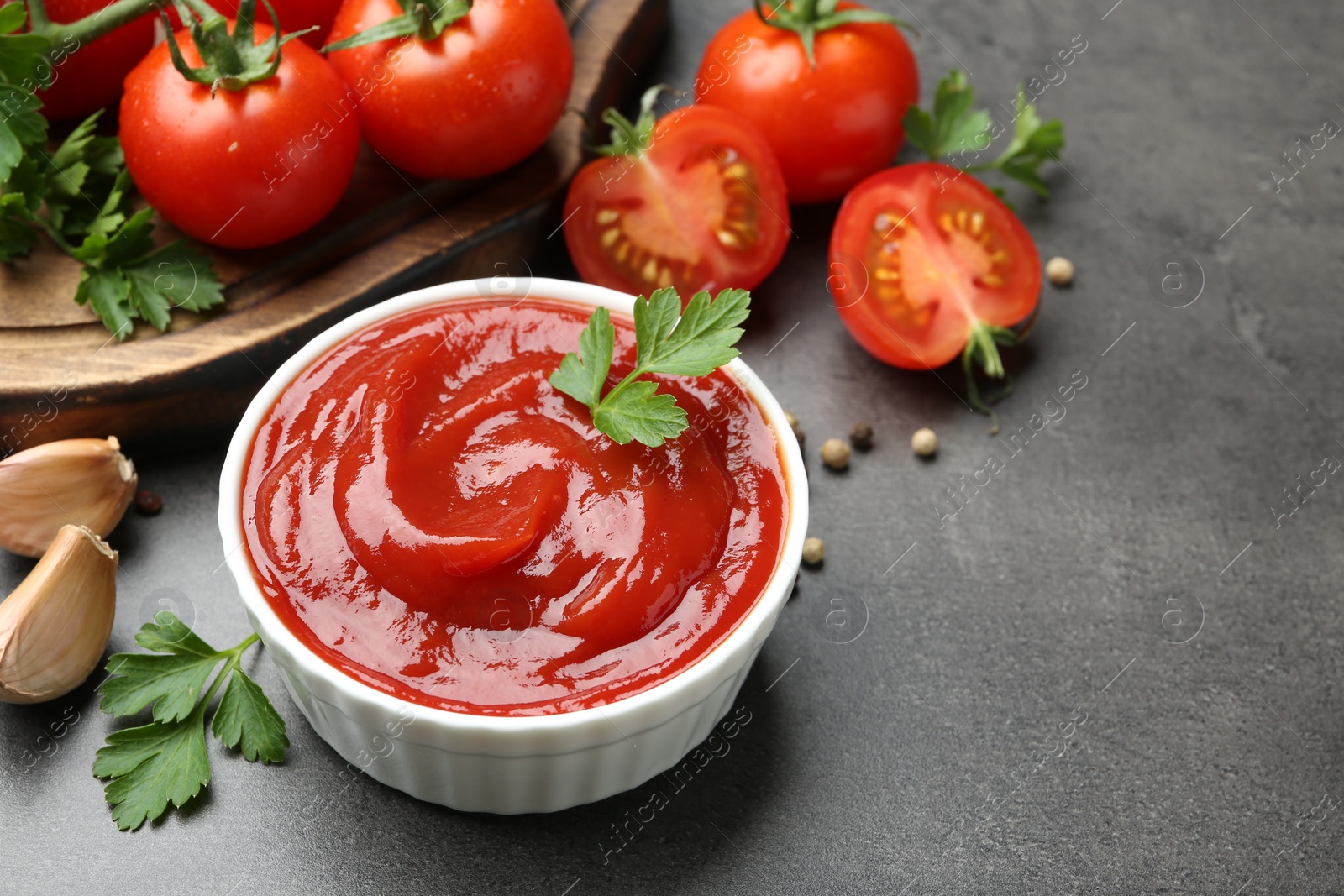 Photo of Delicious ketchup in bowl, tomatoes, parsley and garlic on grey table, closeup