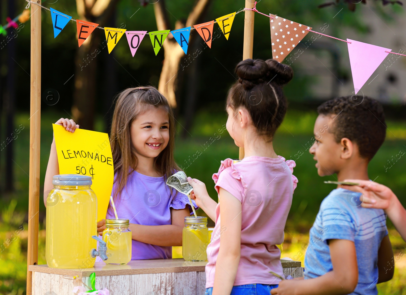 Photo of Little girl selling natural lemonade to kids in park. Summer refreshing drink