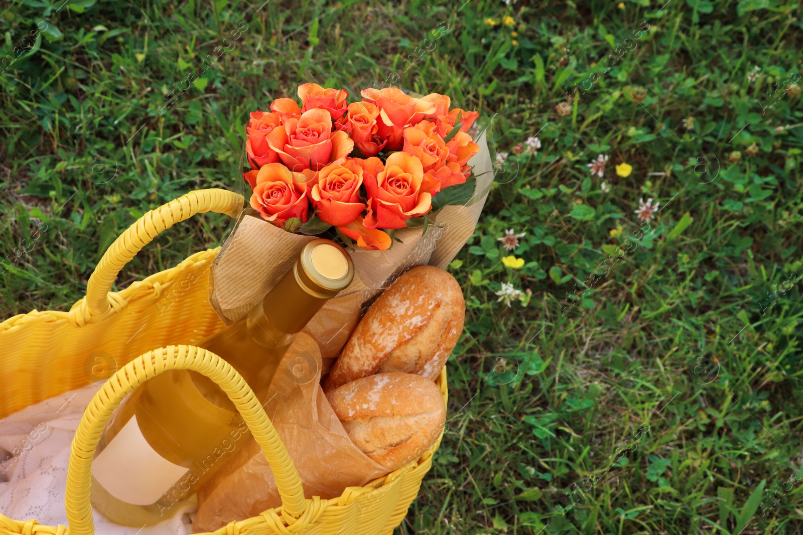 Photo of Yellow wicker bag with beautiful roses, bottle of wine and baguettes on green grass outdoors, above view. Space for text