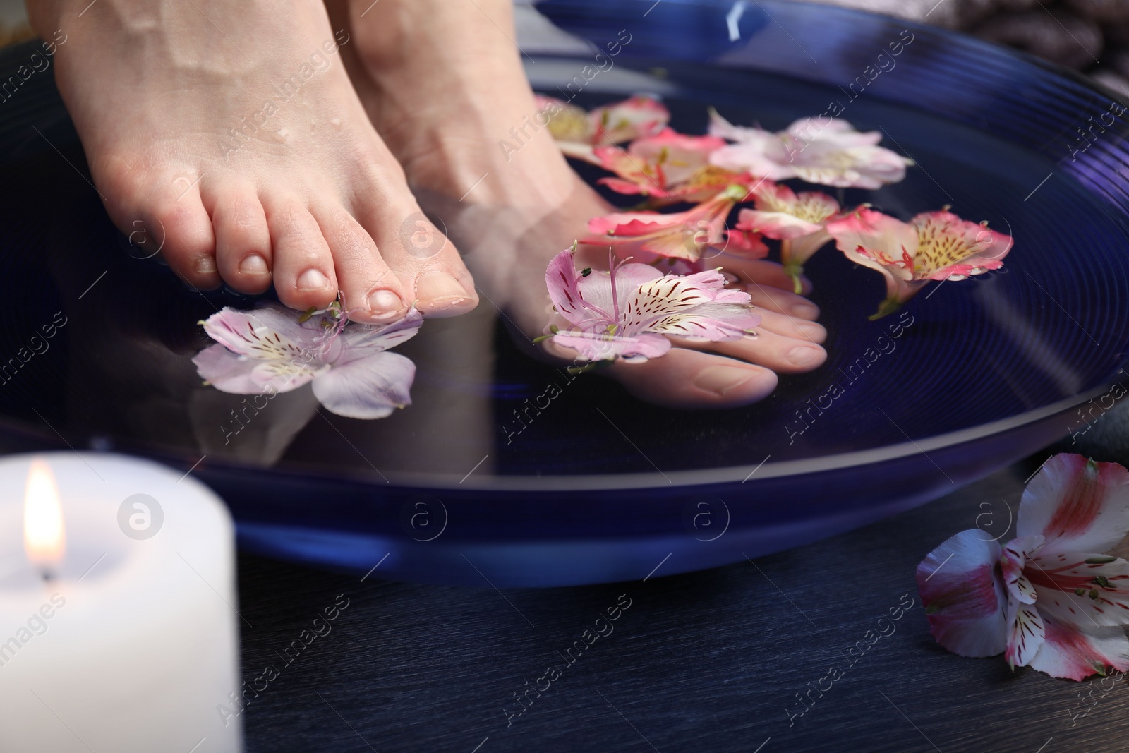 Photo of Woman soaking her feet in bowl with water and flowers on wooden floor, closeup. Spa treatment