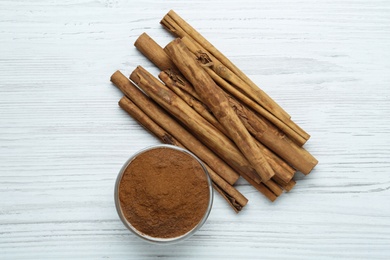 Photo of Aromatic cinnamon powder in bowl near sticks on white wooden table, flat lay