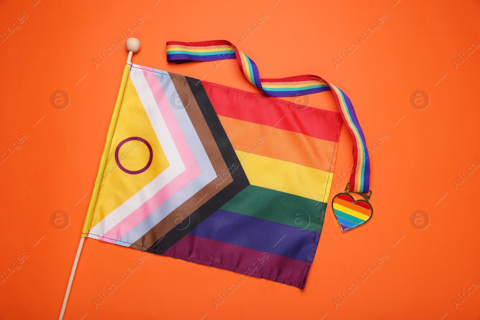 Photo of Bright progress flag and rainbow ribbon with heart shaped pendant on orange background, flat lay. LGBT pride