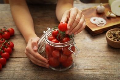 Woman putting tomatoes into glass jar at wooden kitchen table, closeup. Pickling vegetables