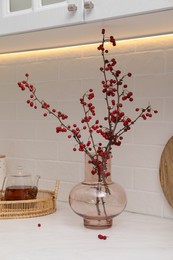 Hawthorn branches with red berries in vase and aromatic tea on white table indoors