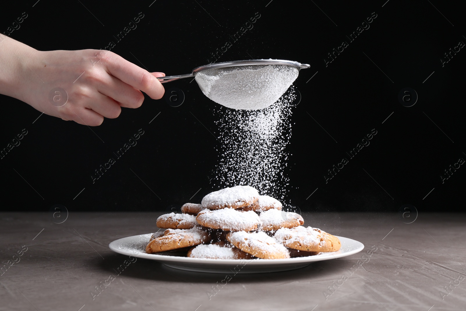 Photo of Woman with sieve sprinkling powdered sugar onto cookies at grey textured table, closeup
