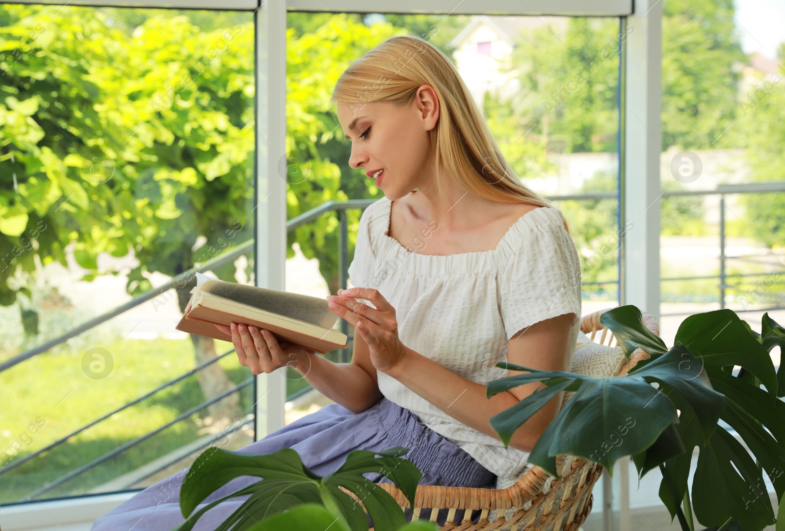 Photo of Young woman reading book in armchair at indoor terrace