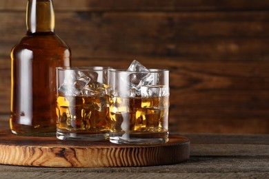 Whiskey with ice cubes in glasses and bottle on wooden table, closeup. Space for text