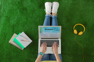 Woman holding laptop with open travel blogger site on artificial grass, top view