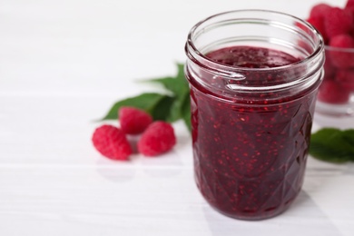 Photo of Delicious jam in glass jar and fresh raspberries on white wooden table. Space for text