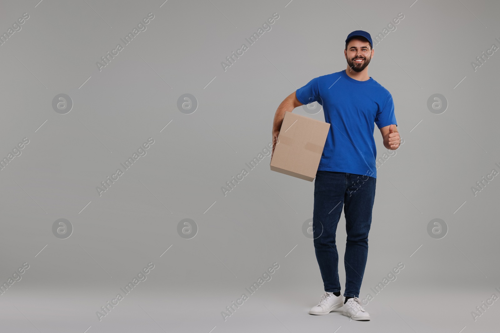 Photo of Happy young courier with parcel showing thumb up on grey background, space for text