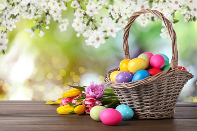 Image of Colorful Easter eggs in wicker basket and tulips on wooden table outdoors, space for text