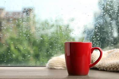 Photo of Cup of hot drink and knitted plaid near window on rainy day. Space for text