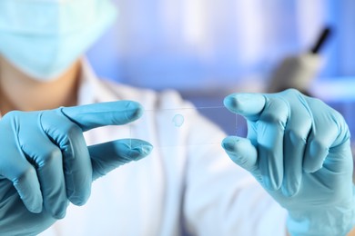 Photo of Scientist holding microscope slide with light blue sample in laboratory, closeup