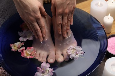 Photo of Woman soaking her feet in bowl with water and flowers on floor, closeup. Spa treatment