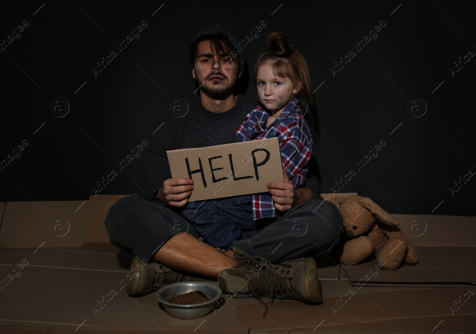 Photo of Poor father and daughter with HELP sign on floor near dark wall