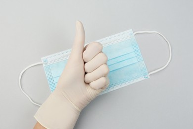 Photo of Doctor in medical gloves showing thumb up gesture near protective mask on light grey background, top view