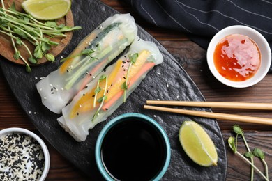 Photo of Tasty spring rolls, sauces, lime, sesame seeds and microgreens on wooden table, flat lay