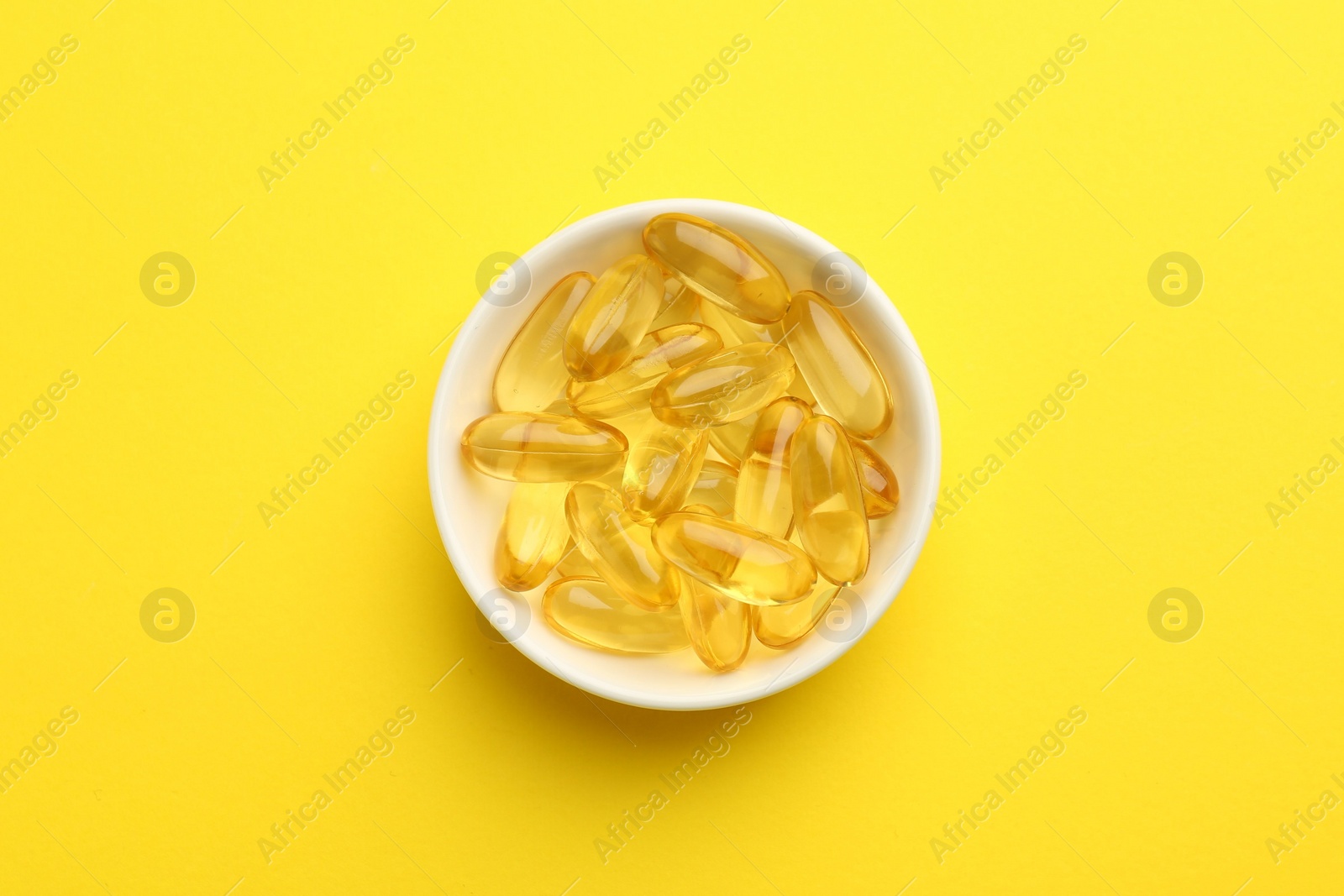 Photo of Vitamin capsules in bowl on yellow background, top view