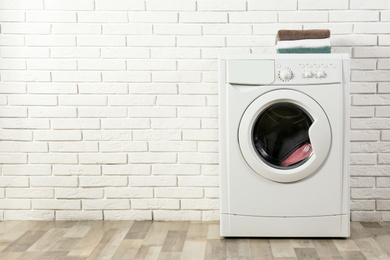 Modern washing machine with laundry and stack of towels near white brick wall. Space for text