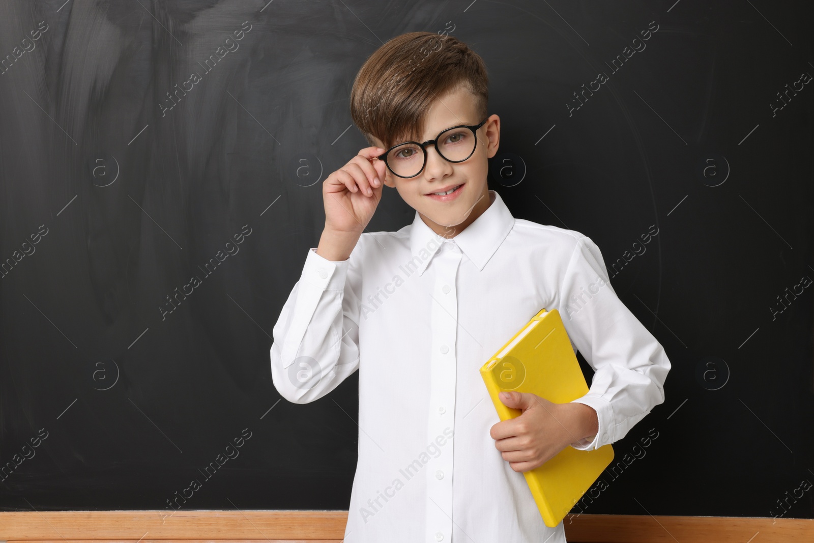 Photo of Cute schoolboy in glasses with book near chalkboard, space for text