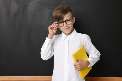 Cute schoolboy in glasses with book near chalkboard, space for text