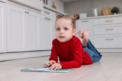 Photo of Cute little girl with book on warm floor in kitchen, space for text. Heating system