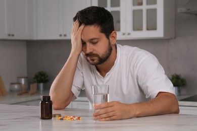 Photo of Depressed man with glass of water and antidepressant pills at table in kitchen