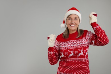 Photo of Happy senior woman in Christmas sweater, Santa hat and knitted mittens on grey background. Space for text