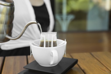 Photo of Woman pouring hot water into cup with drip coffee bag at wooden table, closeup
